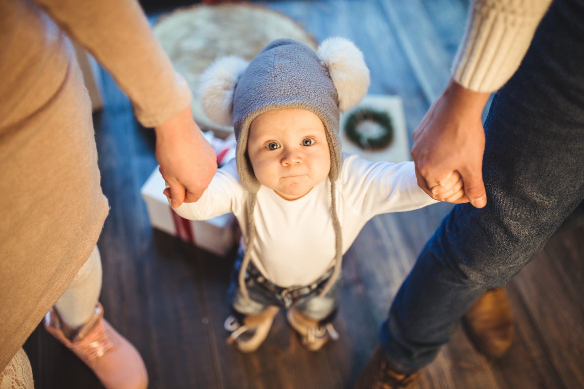 Funny little baby boy 1 year old learning walk home in winter in a decorated New Year house. Young family dad and mom hold by the hands of his son in the loft interior wooden floor near the window