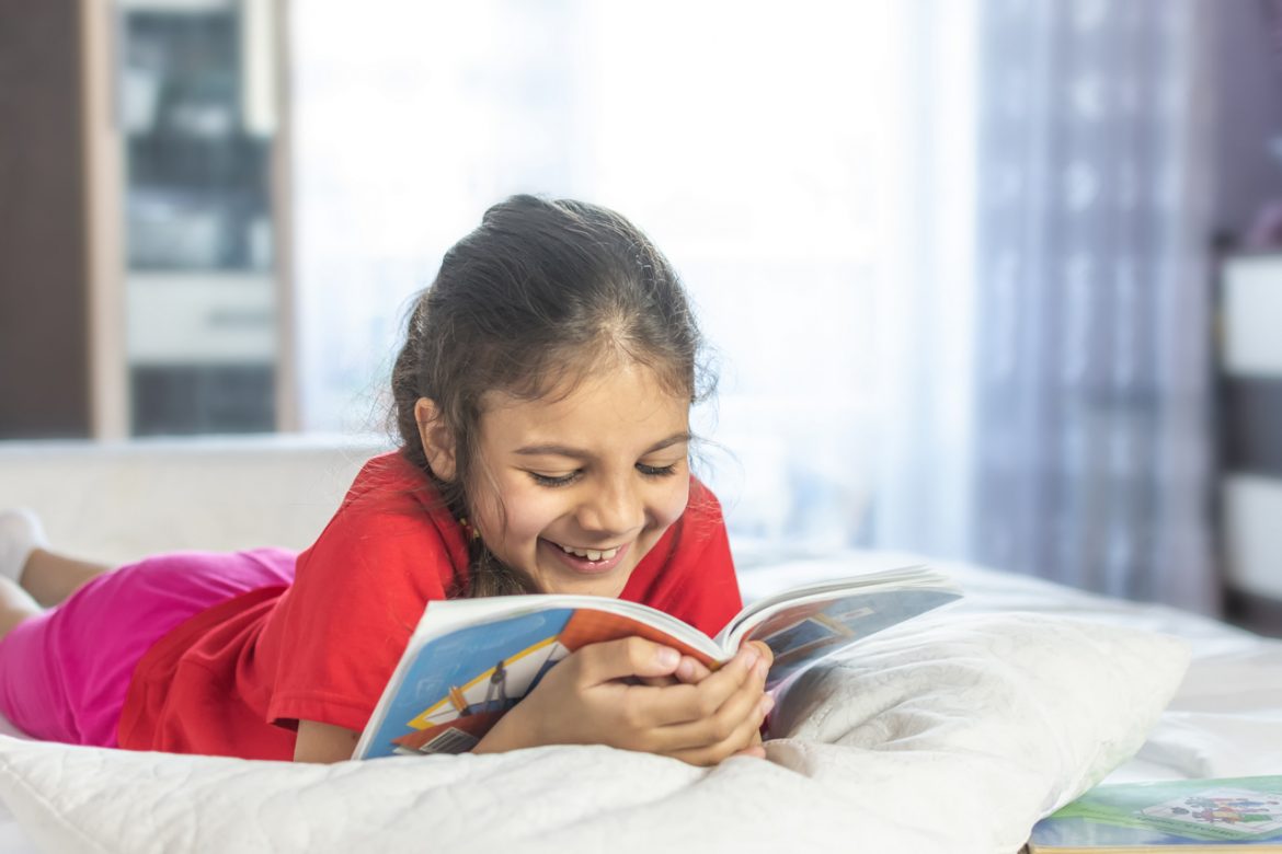 Beautiful girl reading book and smiling. Girl wearing red T-shirt, studying at home with books and doing school homework. Smiling girl.